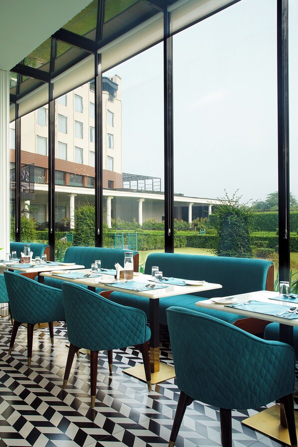 A restaurant with teal upholstered seating, a tiled floor, and large windows offering a view of a building and greenery.