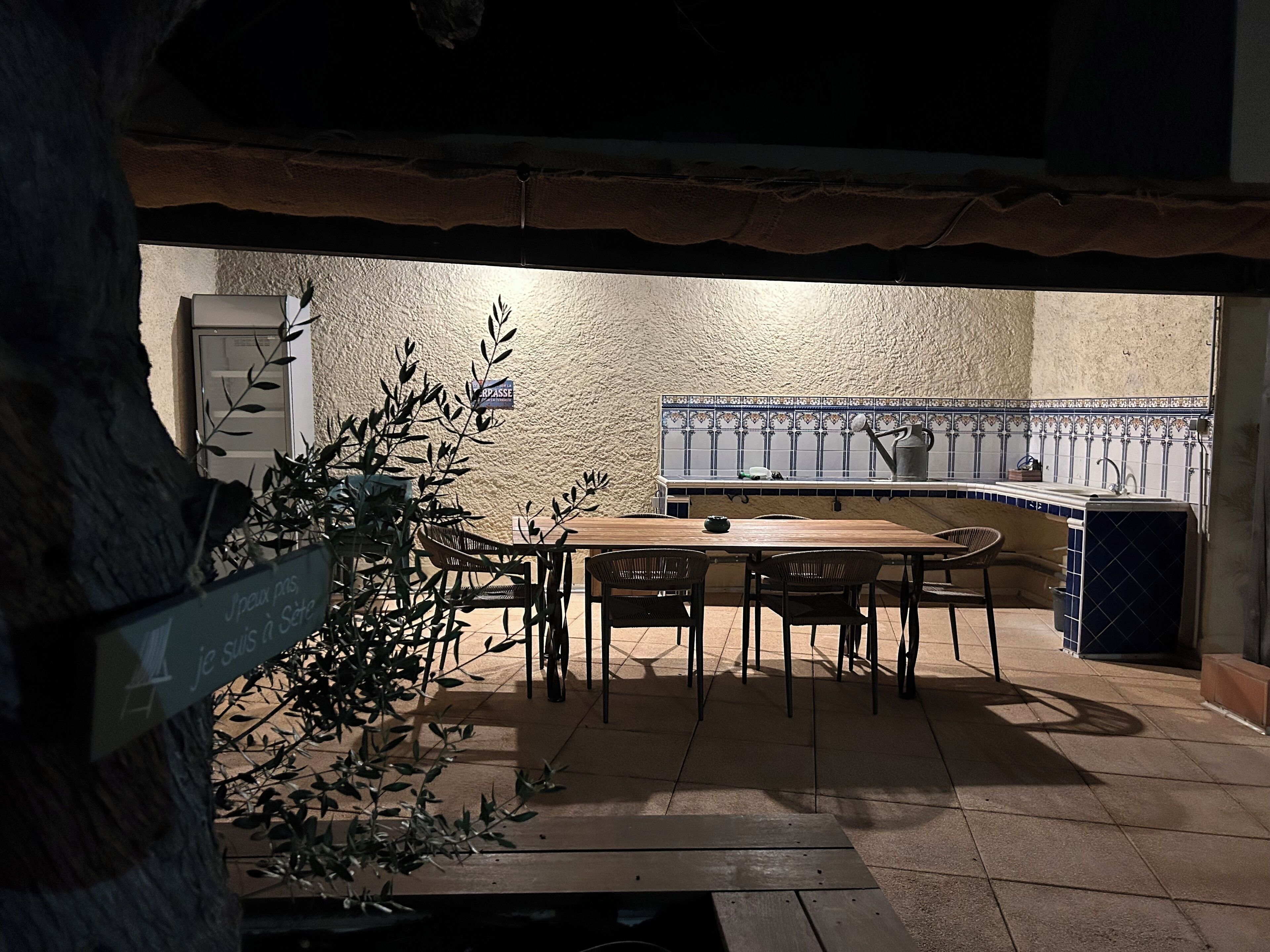 A dining area with a table and chairs, a kitchen with blue and white tiles, and a sign in French.
