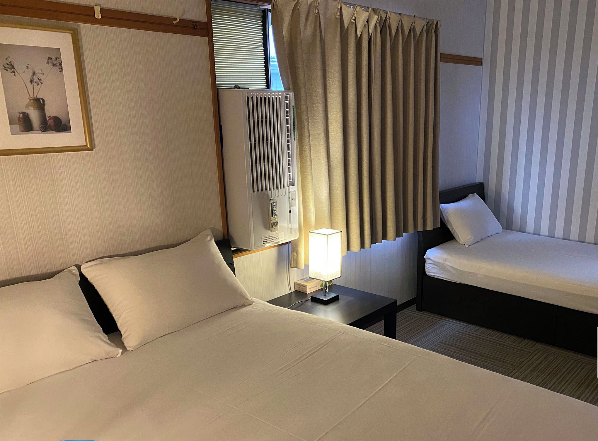 Camera d'albergo con un letto grande, un letto più piccolo, un comodino con una lampada, una finestra con tende e un quadro appeso al muro.