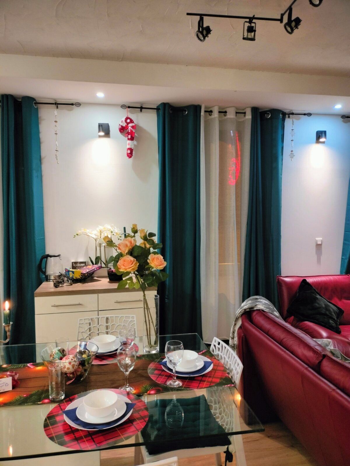 Uma área de refeições com uma mesa de vidro posta para dois, um sofá vermelho e cortinas verde-água.