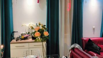 Uma área de refeições com uma mesa de vidro posta para dois, um sofá vermelho e cortinas verde-água.