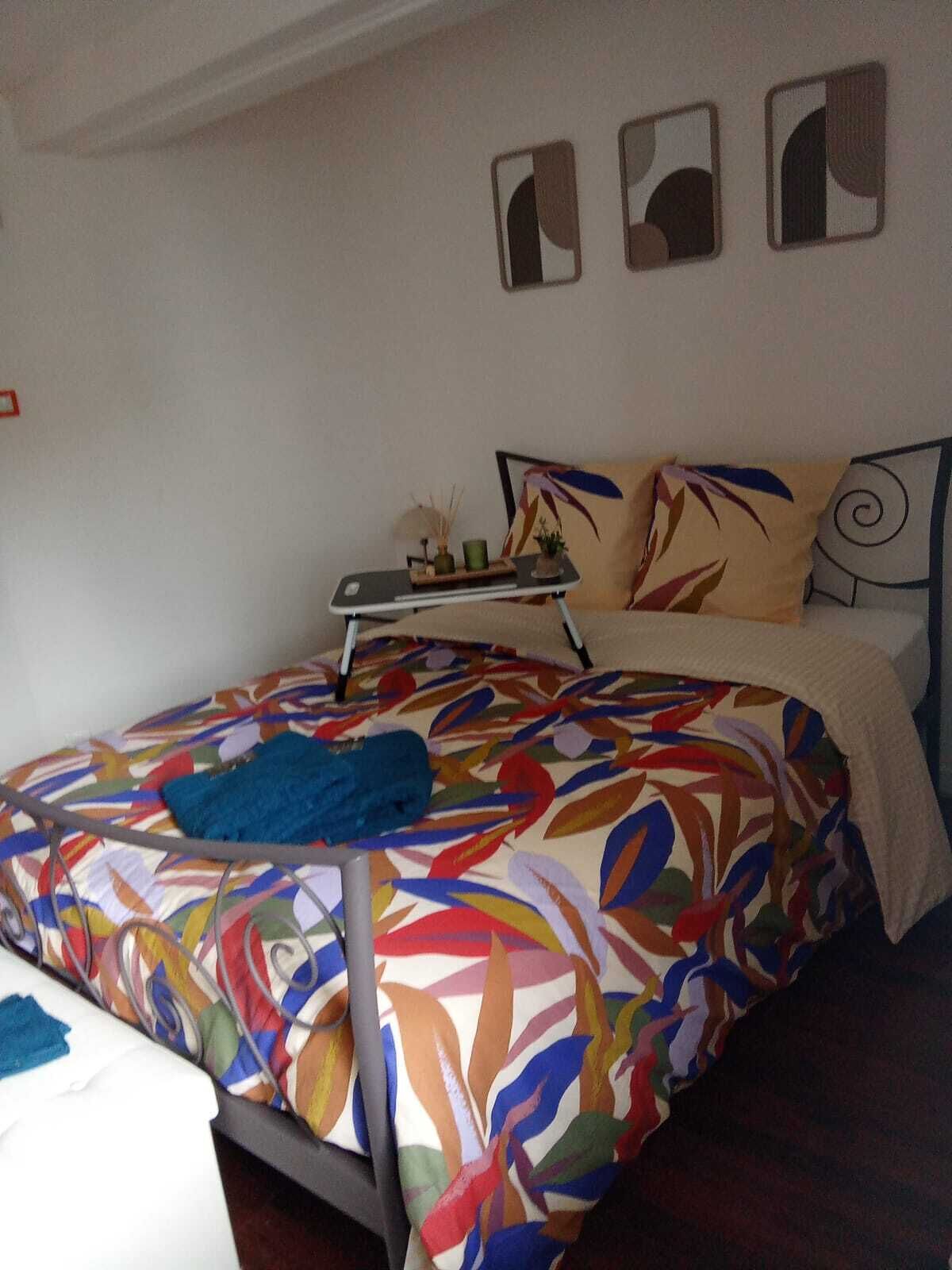 Un lit recouvert d’une couvre-lit aux motifs floraux colorés, une table de chevet avec une lampe et trois œuvres d’art accrochées au mur.