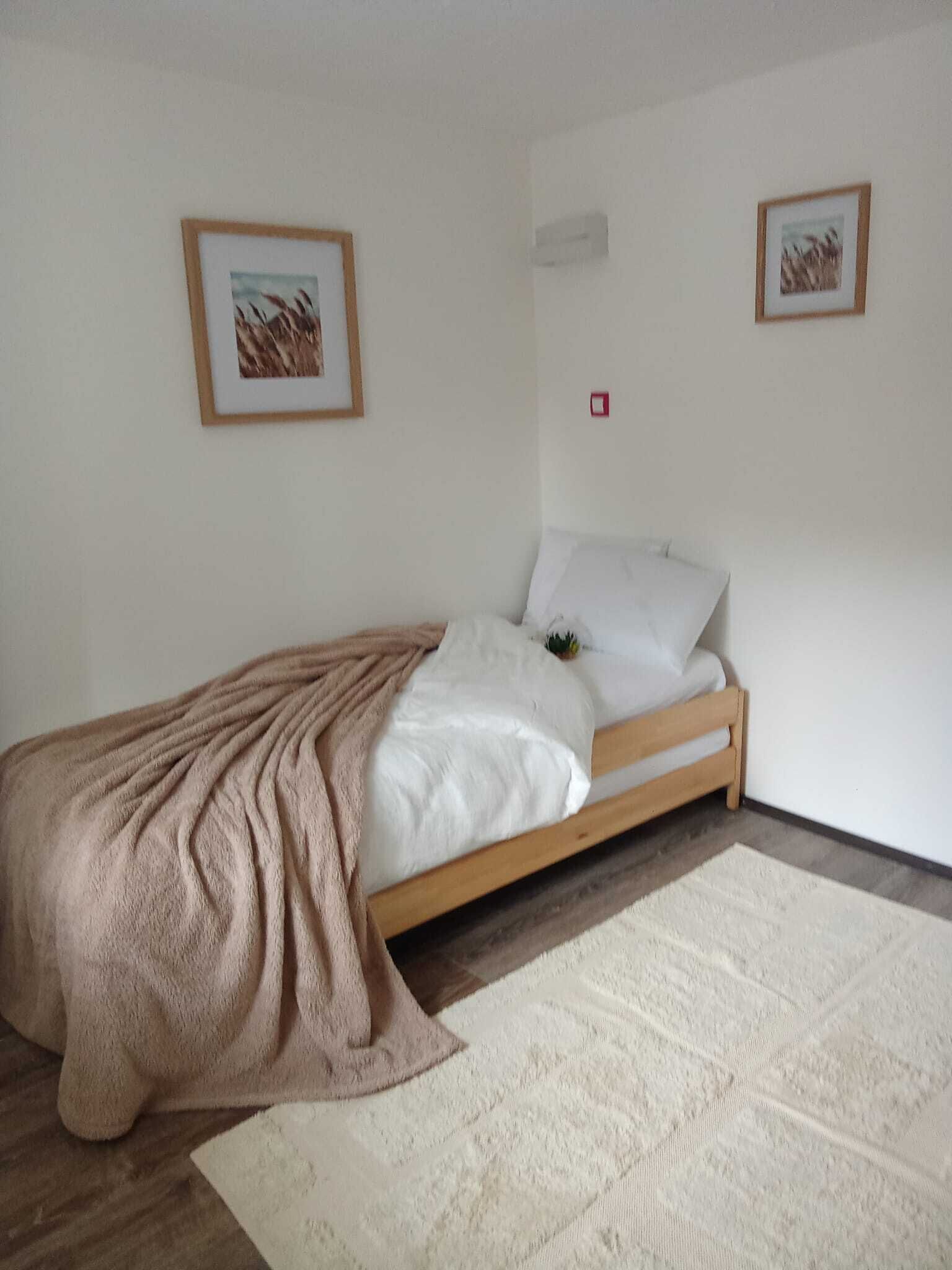 Een eenpersoonskamer met een houten bedframe, witte beddengoed, een beige deken en een witte vloerkleed. Er hangen twee ingelijste foto’s aan de muur.