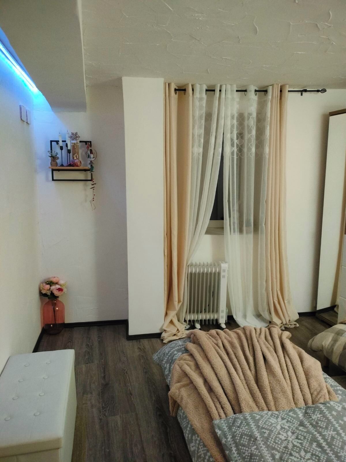 A bedroom with a bed, a bedside table, a radiator, and a shelf with decorative items.