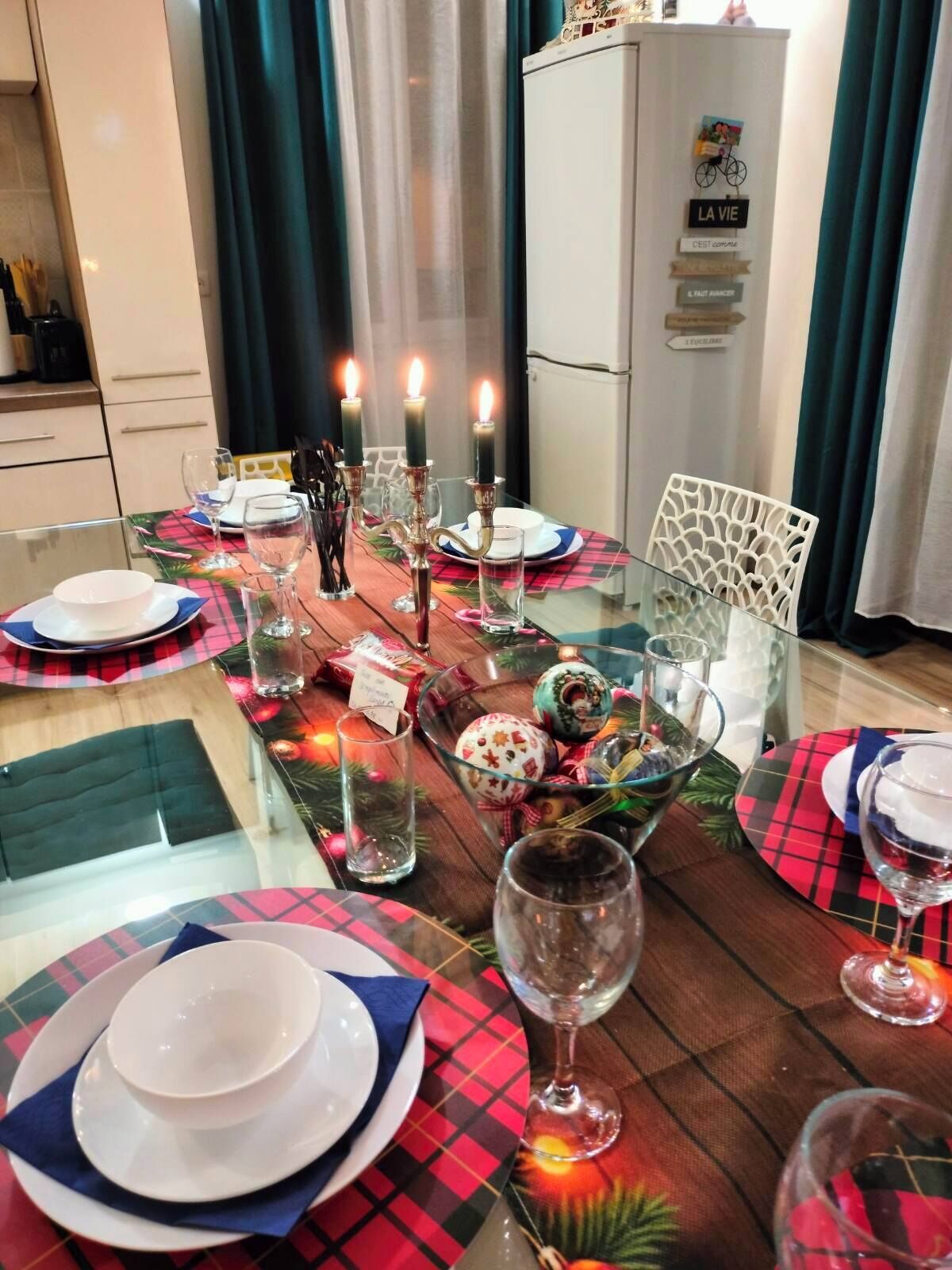 Une table à manger dressée avec des assiettes, des verres et des bougies, entourée de rideaux turquoise et d’un réfrigérateur sur lequel est apposée une pancarte.