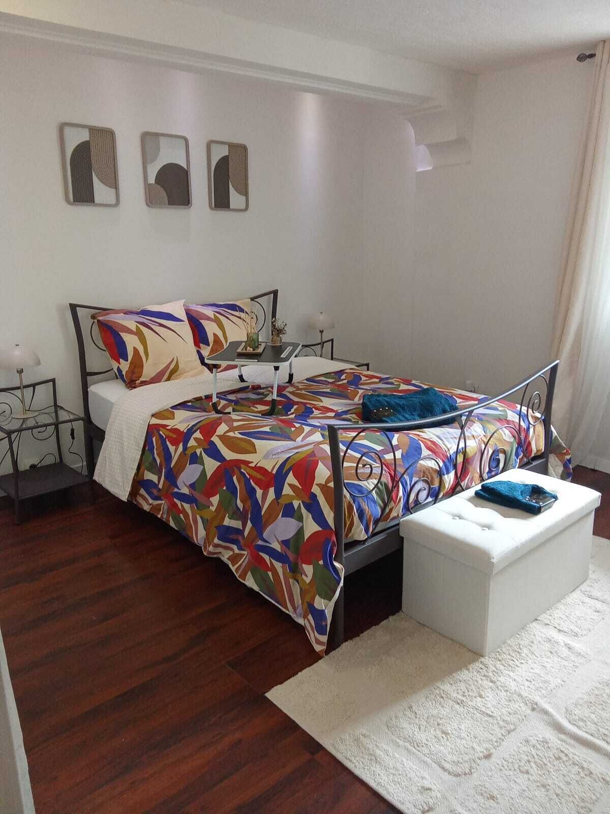 Quarto com cama, mesa de cabeceira, otomano branco e parede com três obras de arte emolduradas.