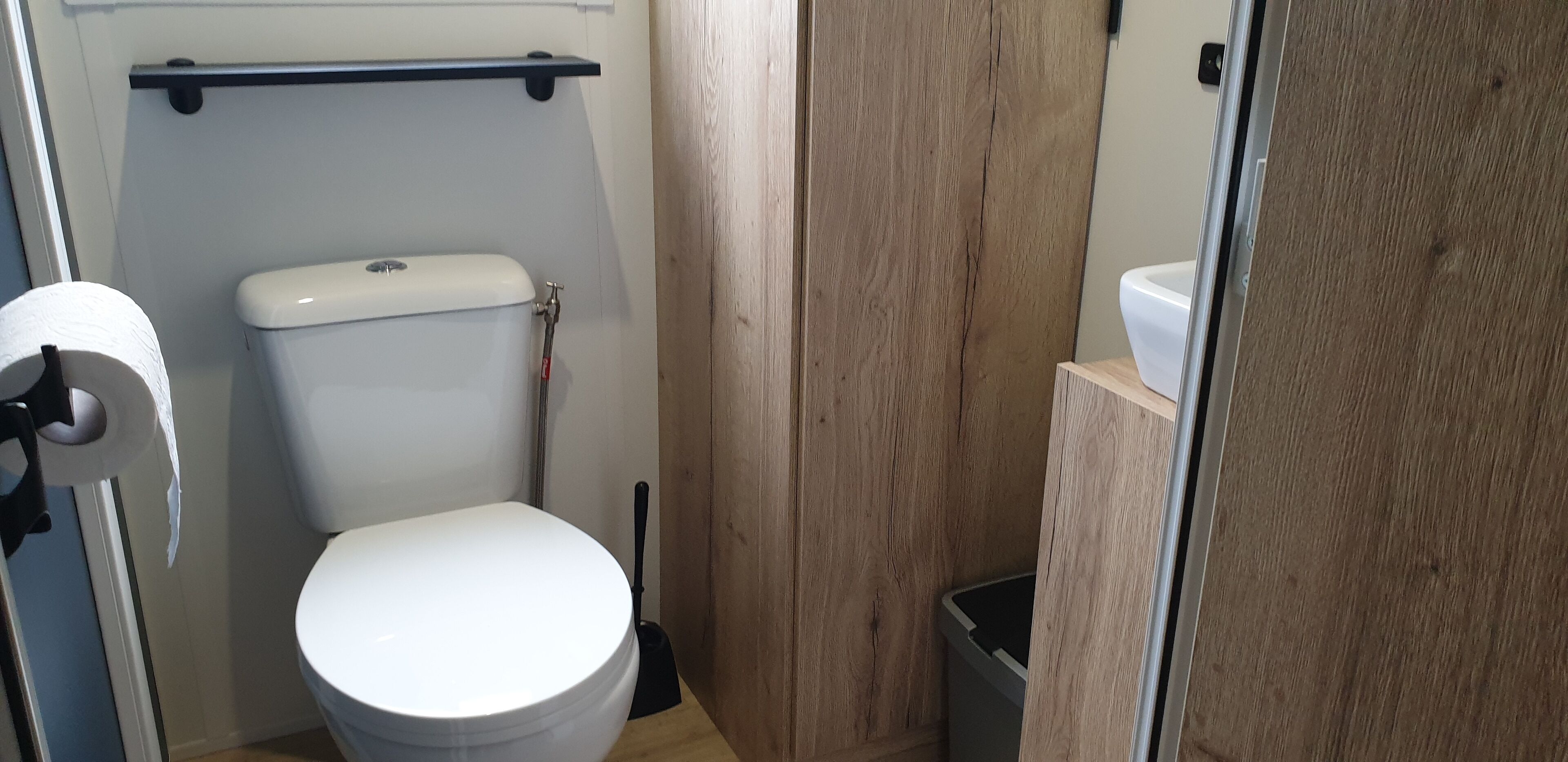 A compact bathroom with a toilet, a toilet paper holder, a black trash bin, and a wooden wall.