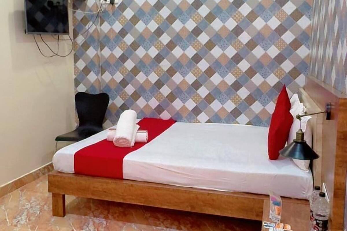 Una cama individual con una colcha roja y blanca, una mesita de noche de madera con una botella, un control remoto y un paño rojo. Un equipo de aire acondicionado está montado en la pared sobre la cama.