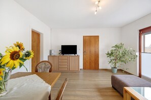 Een kamer met een houten eettafel, een televisie op een houten kast, een bank en een kamerplant.