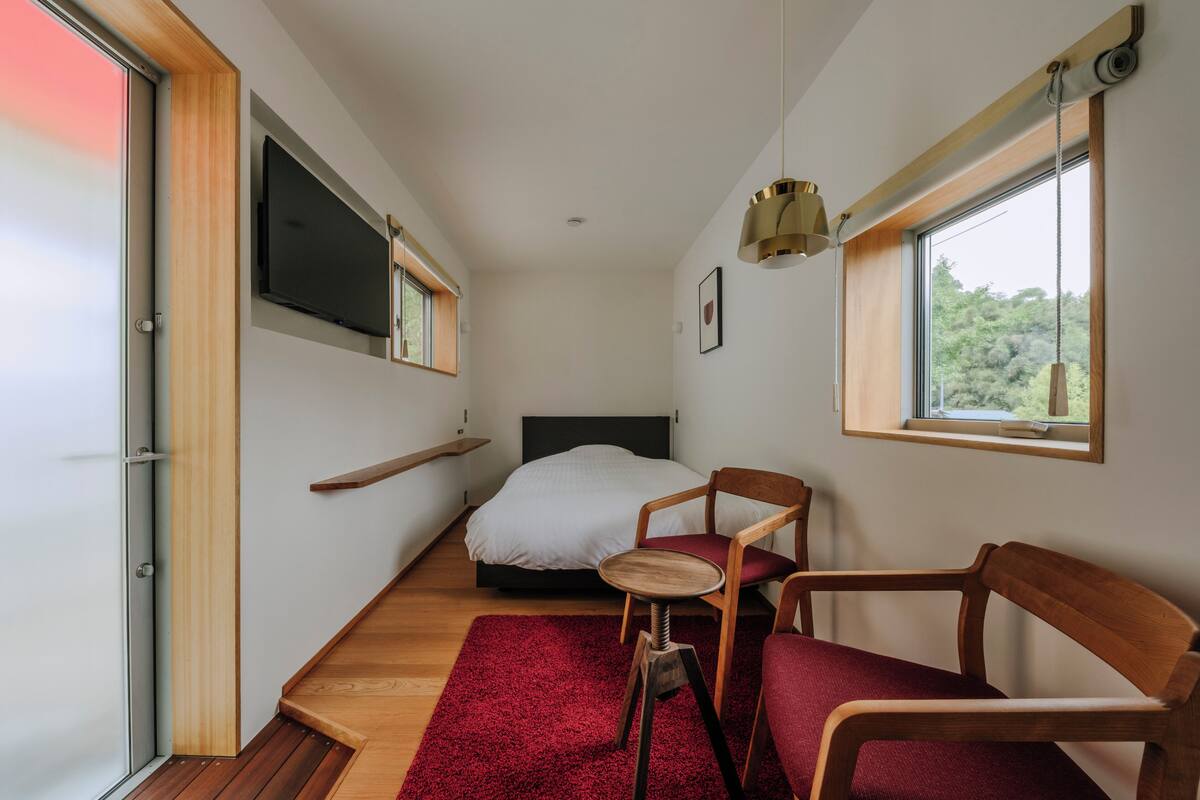Een compacte slaapkamer met een bed, een stoel, een klein tafeltje, een televisie en een raam met uitzicht op groen.