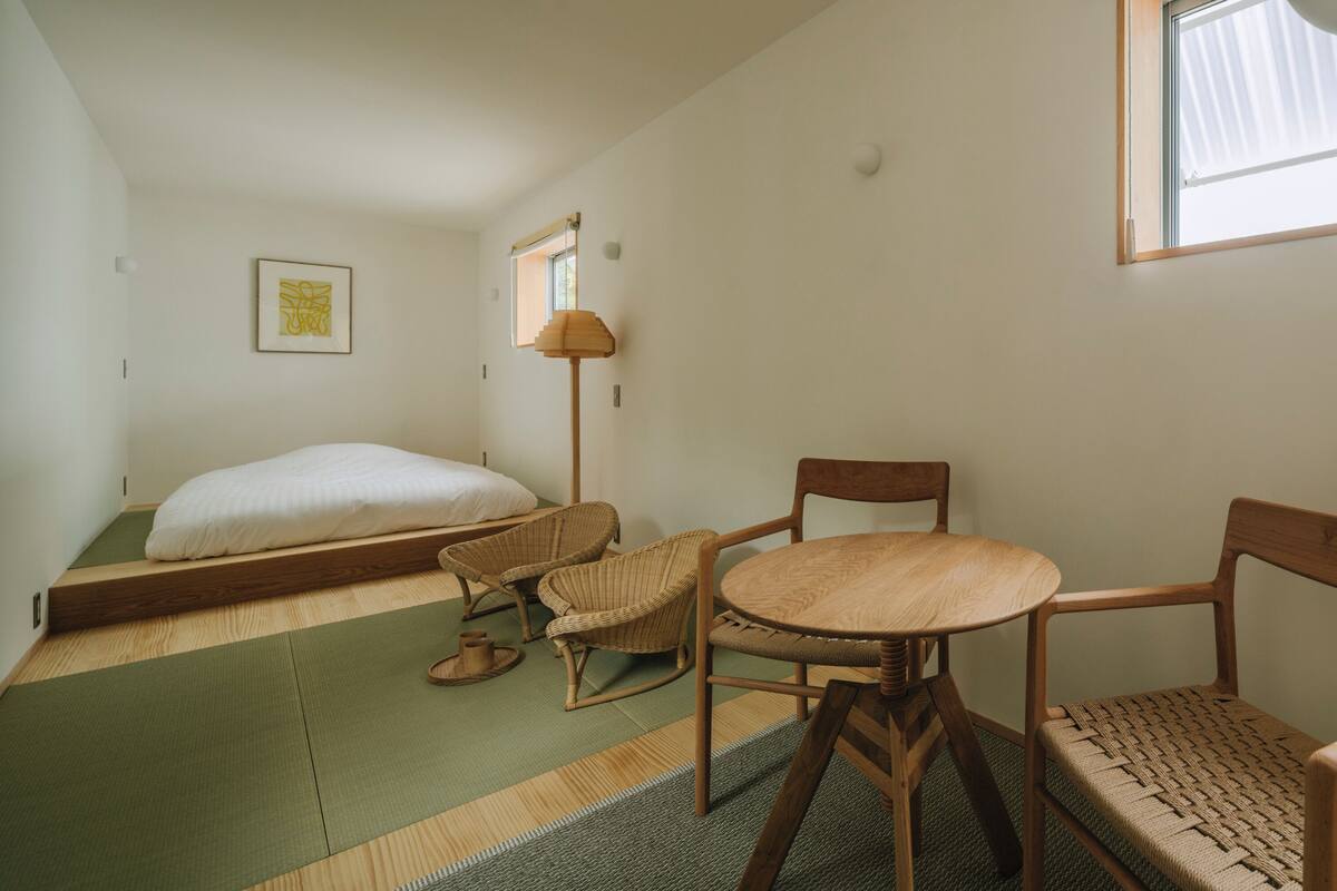 Een kamer met een bed, twee stoelen, een tafeltje, een vloerlamp, een raam en een fotokader aan de muur.