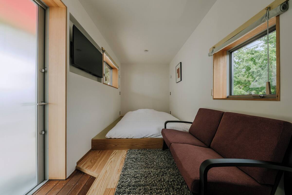 Een knus slaapkamer met een bed, een slaapbank, een aan de muur bevestigde televisie en een raam met uitzicht op groen.