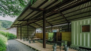A covered outdoor seating area with tables and chairs, a vending machine, and a view of a green building in the background. - NANUKA HOTEL (Kirishima)
