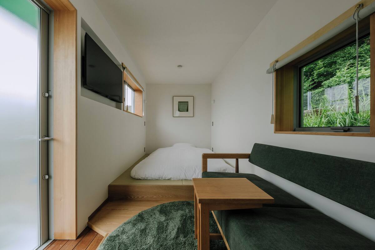 Een compacte kamer met een bed, een groene slaapbank, een klein tafeltje, een aan de muur bevestigde televisie en een raam met uitzicht op groen.
