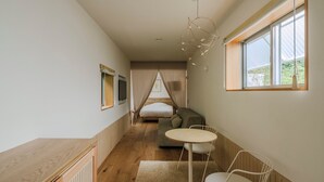 A modern, minimalist room with a wooden floor, a round table with chairs, a sofa, a bed with a canopy, and a large window with a view of greenery. - NANUKA HOTEL (Kirishima)