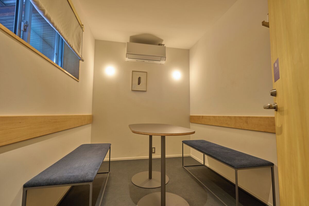 Um espaço pequeno e fechado, com uma mesa redonda e dois bancos, um aparelho de ar condicionado e uma janela com persianas.