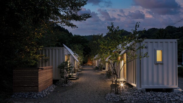 A row of compact, single-story cabins with a gravel path leading through them, surrounded by trees and a wooden fence. - NANUKA HOTEL (Kirishima)