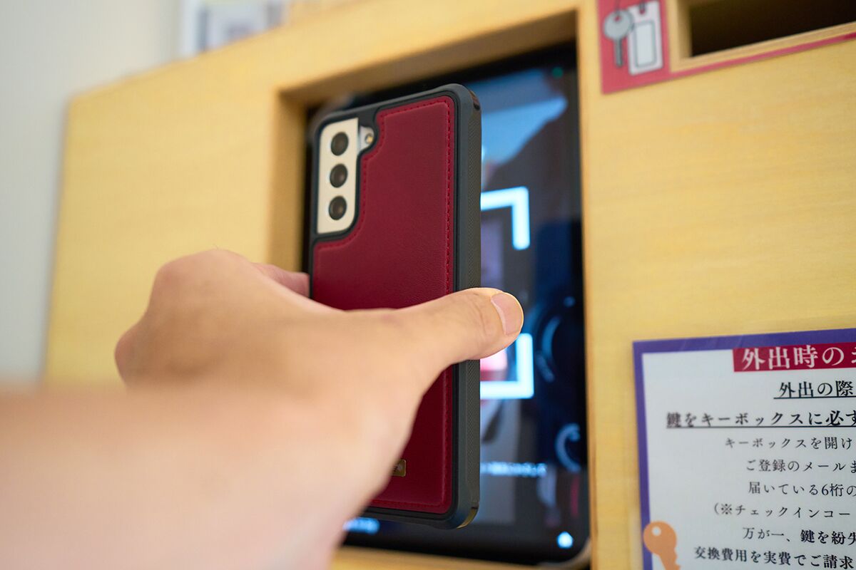 Une main tenant un smartphone avec une coque rouge, placé devant un écran numérique affichant du texte en japonais.