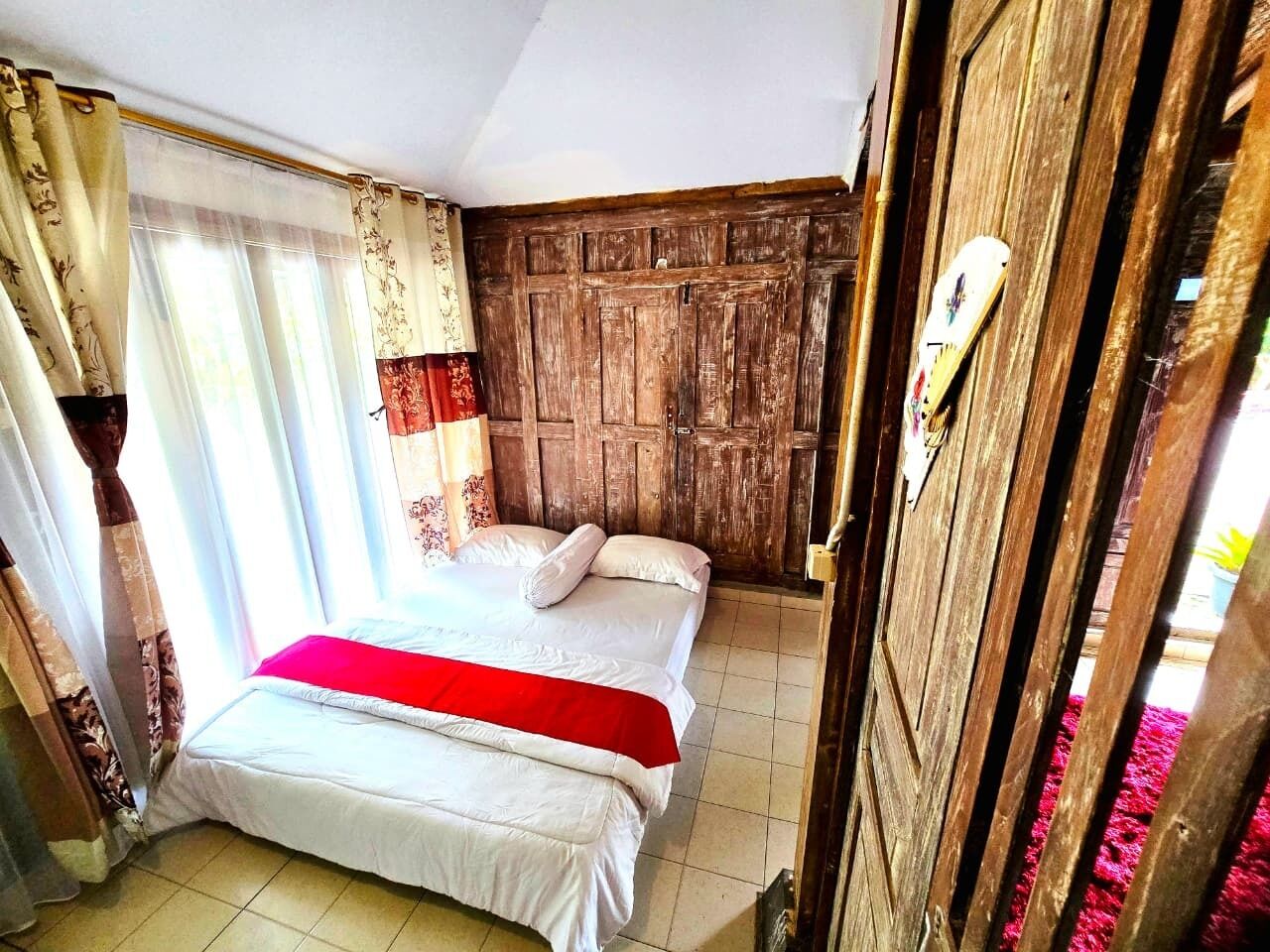 Une pièce avec une porte en bois, un lit recouvert de draps blancs et d’une couverture rouge, et une fenêtre ornée de rideaux légers.
