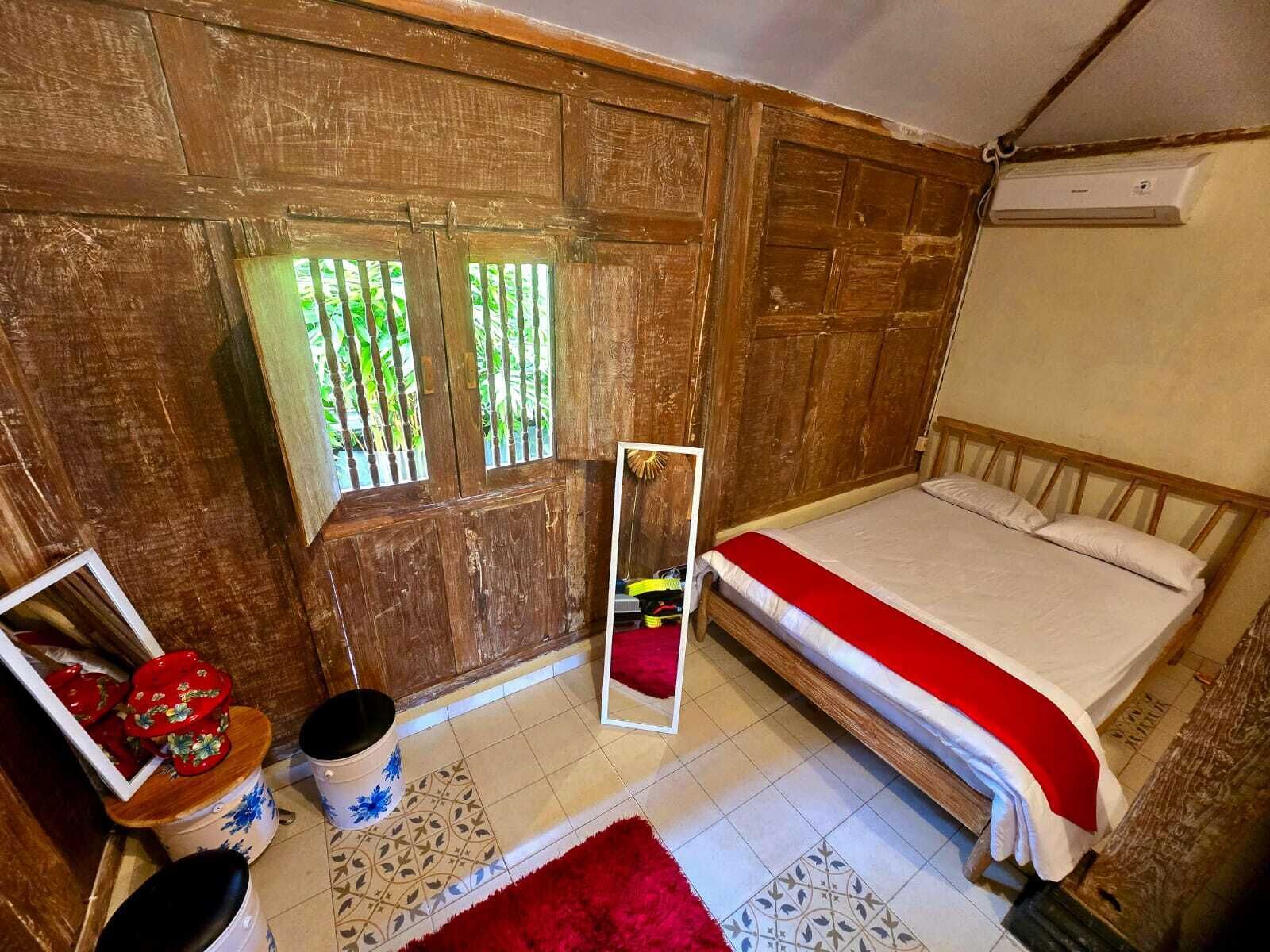 A room with a wooden wall, a bed with a red blanket, a small table with a mirror, and a window with a view of greenery.