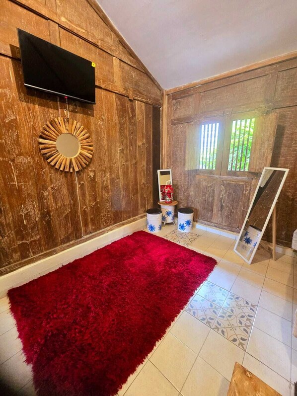Une pièce avec un mur en bois, un tapis rouge, un téléviseur fixé au mur et une fenêtre avec des stores verticaux.