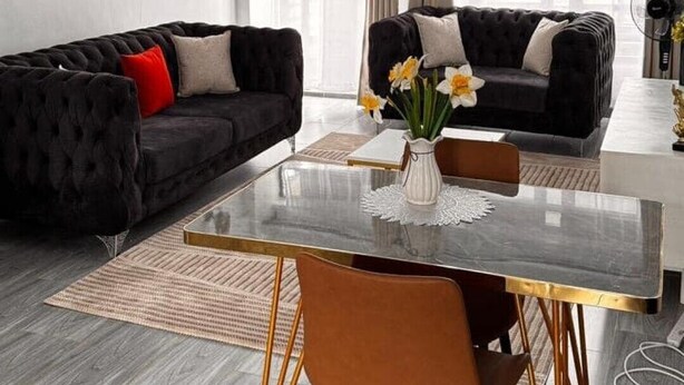 Een moderne woonkamer met een zwarte bank, een eettafel met stoelen en een groot raam met transparante gordijnen.