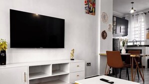 Een moderne woonkamer met een flatscreen televisie bevestigd aan een witte muur, een witte tv-meubel eronder en een klein eethoekje met een tafel en stoelen.