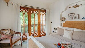 A bedroom with a large bed, a chair, and a balcony with wooden doors and a view of greenery.