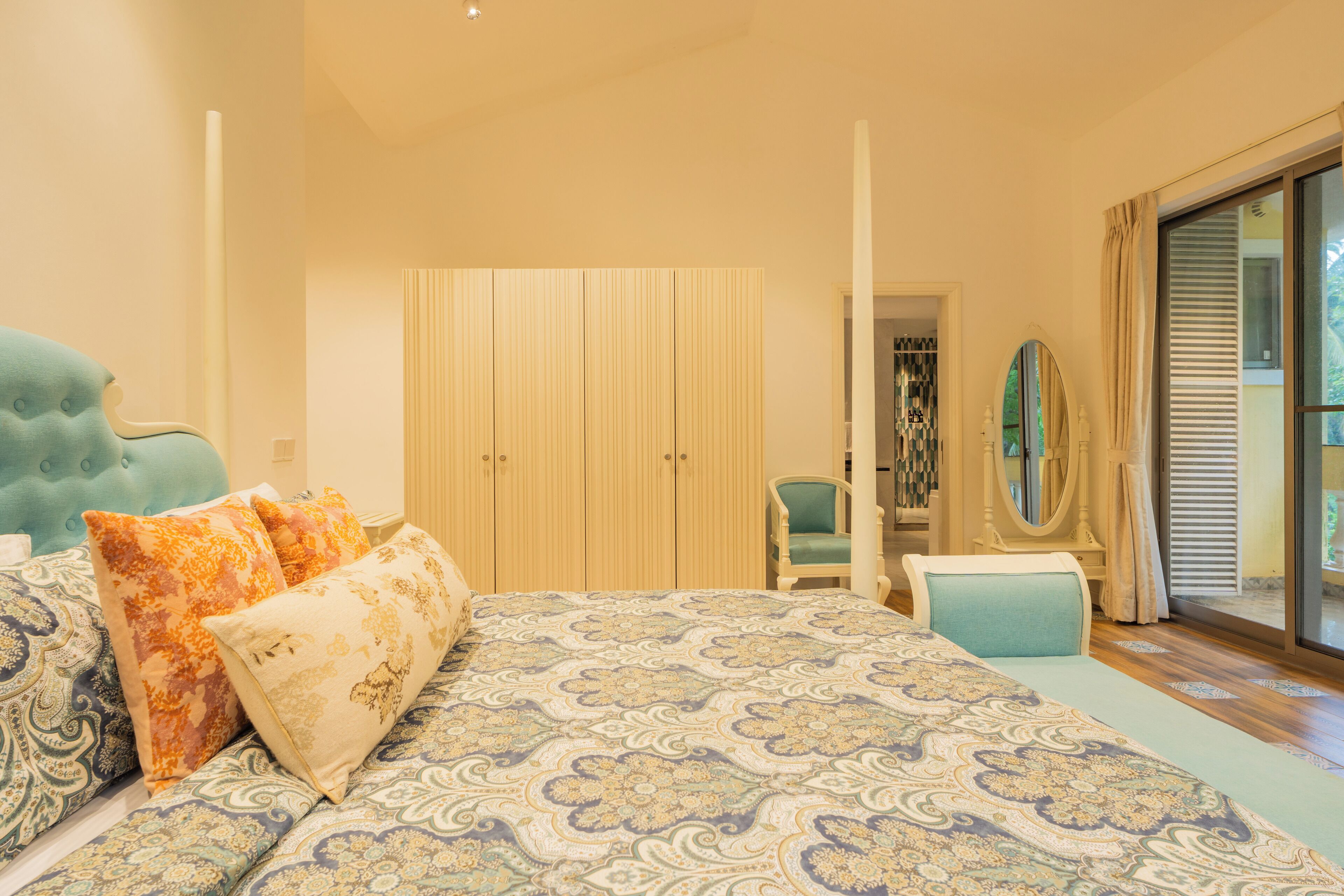 A bedroom with a large bed, a wardrobe, a vanity with a mirror, and a view through a sliding glass door.