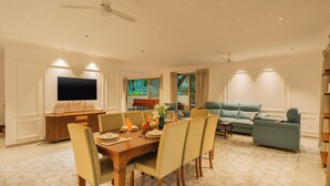 A dining area with a wooden table and chairs, a television mounted on the wall, and a living area with a sofa and a window with curtains.