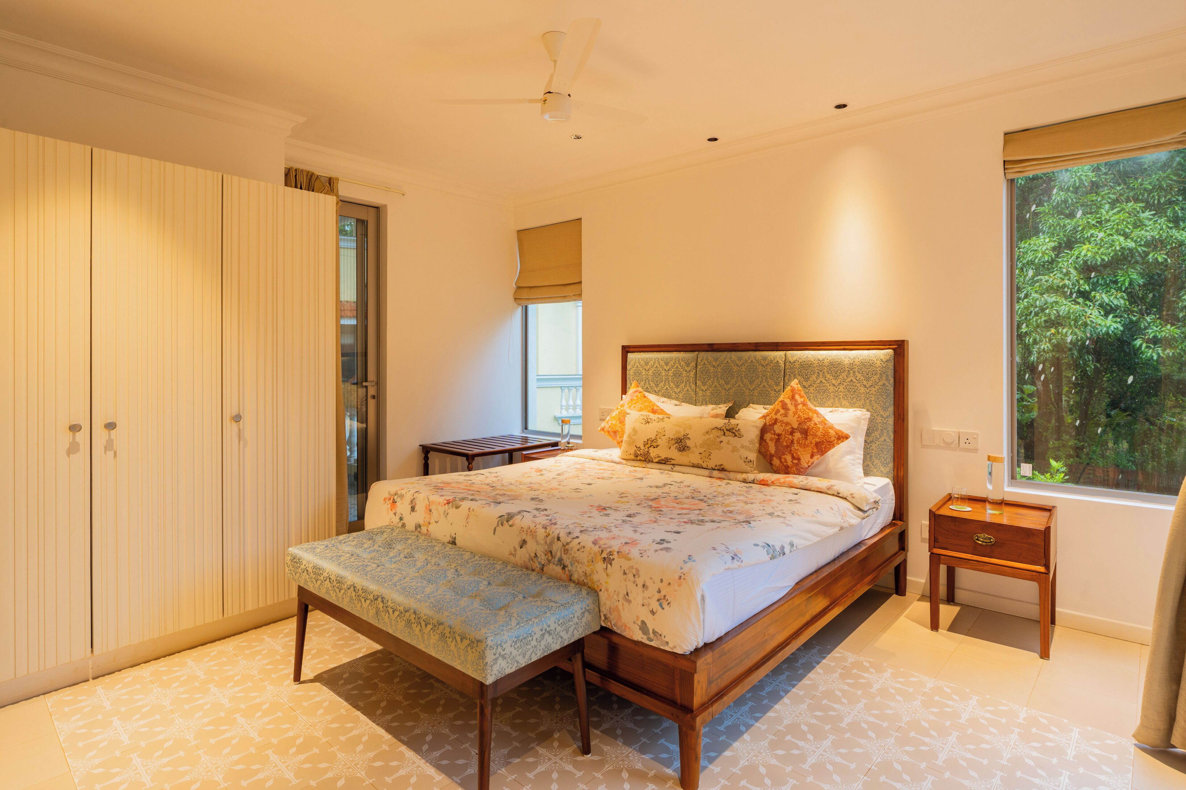 A bedroom with a bed, bench, wardrobe, bedside table, and a window with a view of greenery.