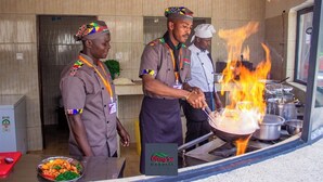 A kitchen with chefs cooking over an open flame, a tiled wall, and a counter with a dish of food. - Oleng'oti Gardens Narok (Narok)