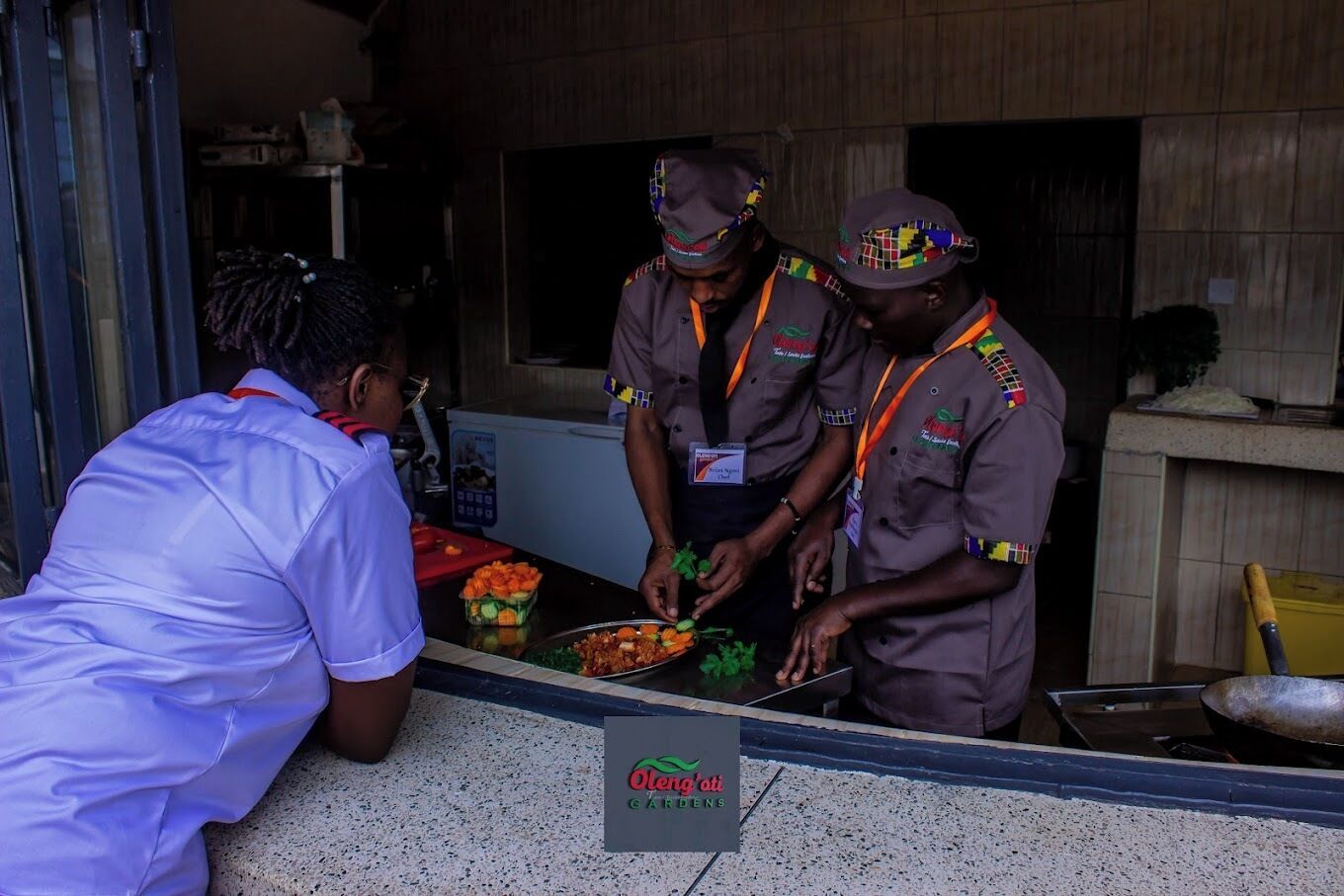 Three individuals in uniform preparing food at a counter with a sign reading "Olen'oti Gardens".