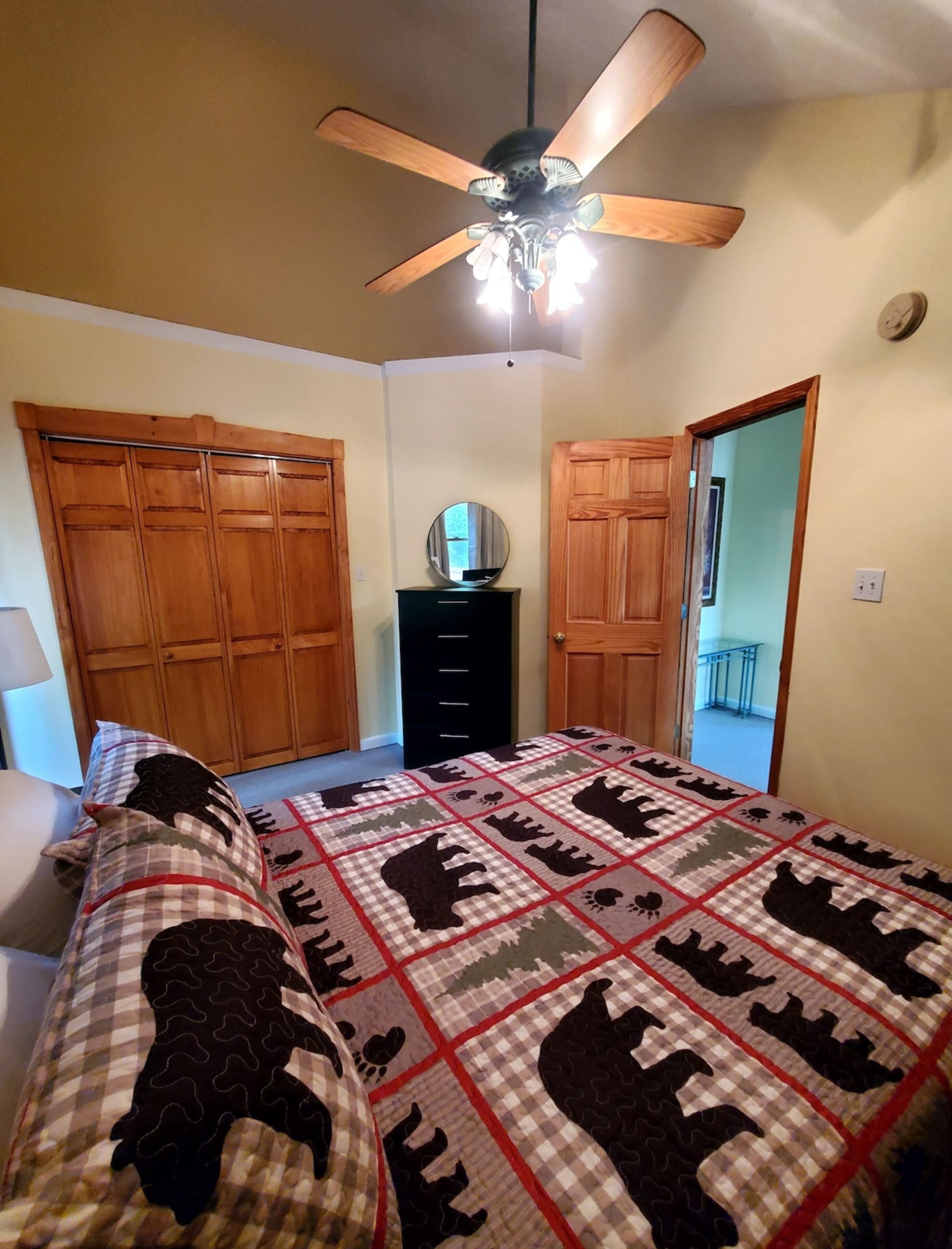 A bedroom with a bed, wooden wardrobe, a dresser, a ceiling fan, and a mirror.