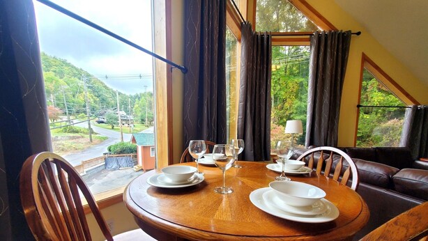 A dining table set with white plates and wine glasses, surrounded by wooden chairs, with a view of a green landscape through large windows.