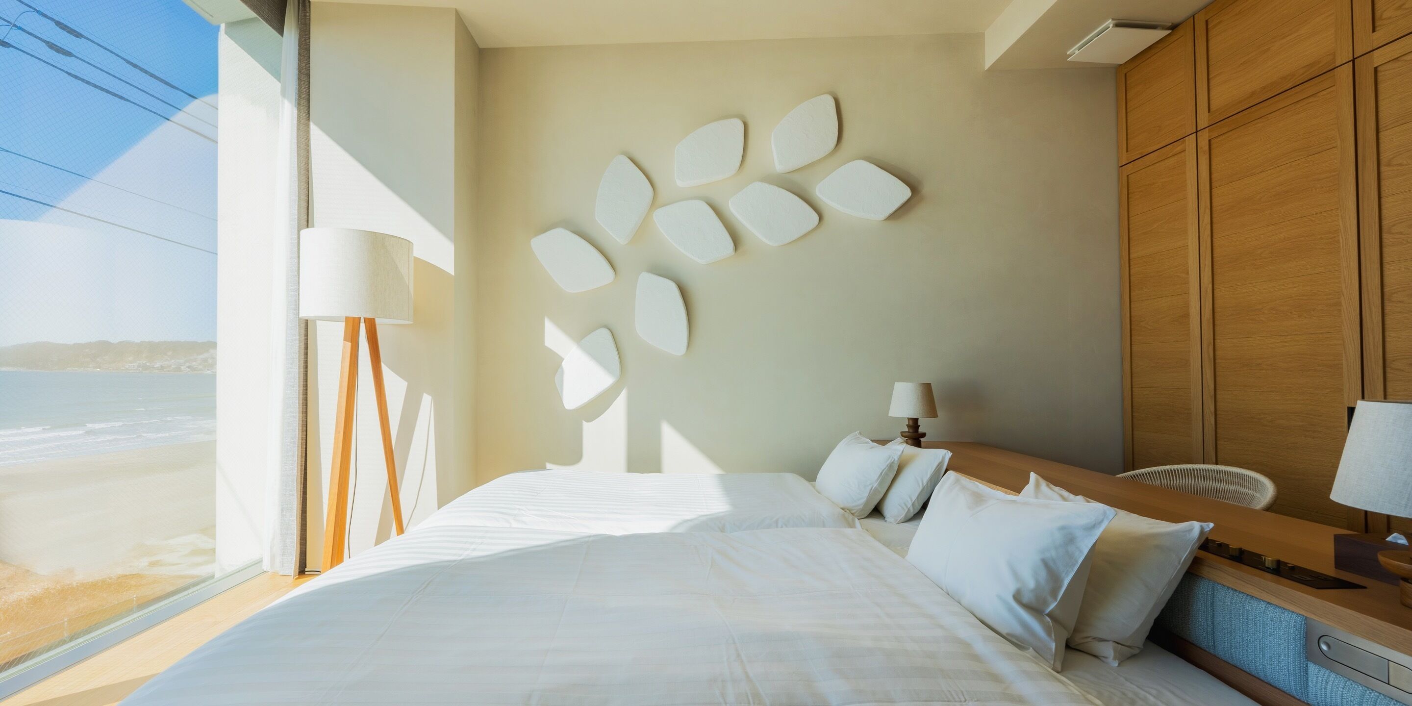 A modern hotel room with a large bed, a wooden headboard, a bedside table with a lamp, and a view of the beach.