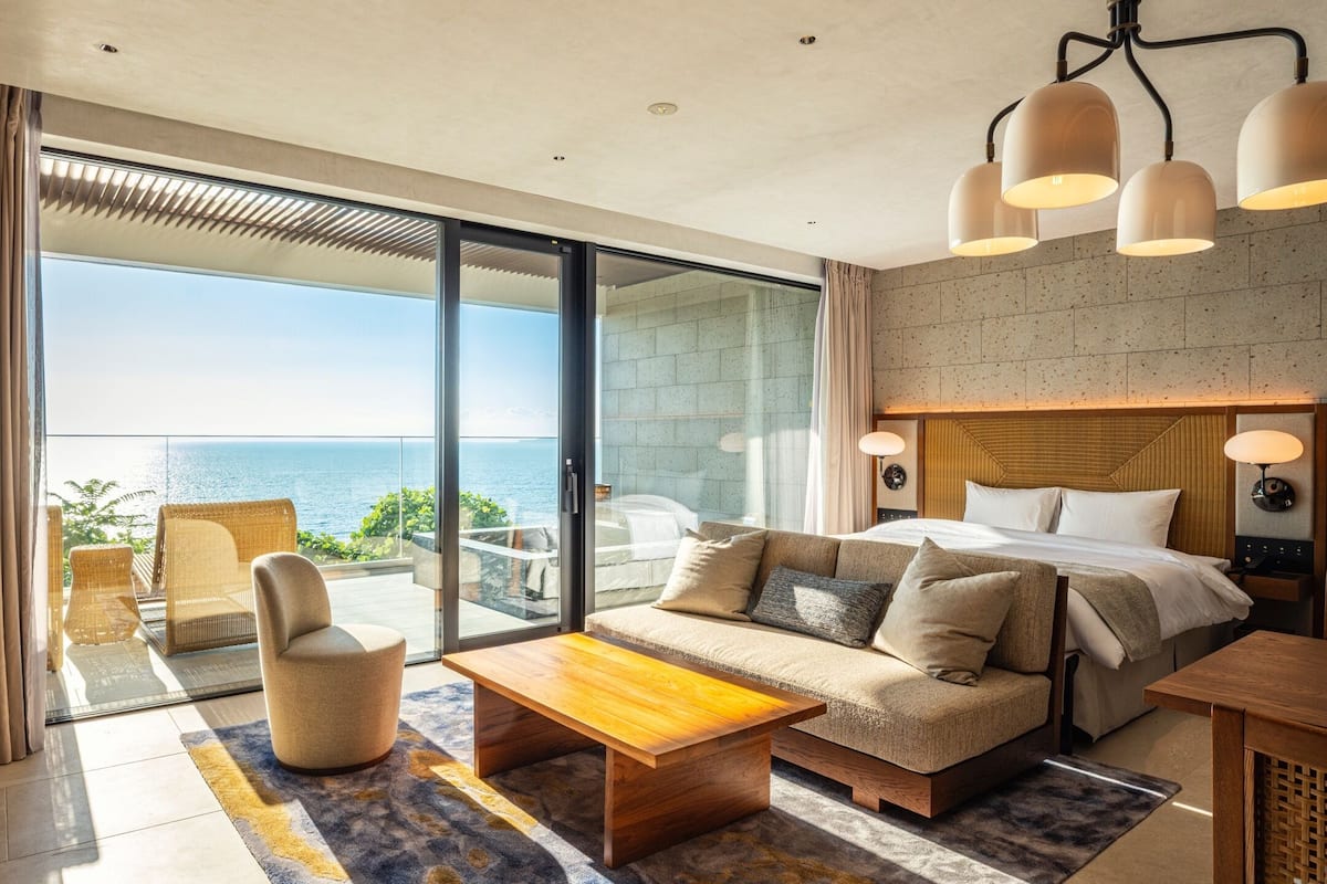 A modern hotel room with a large sliding glass door leading to a balcony with a view of the sea. The room includes a sofa, a bed, a wooden coffee table, and a small table with a lamp.