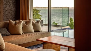A modern living room with a sofa, cushions, and a wooden coffee table. A glass door leads to a balcony with a pool and a view of the sea. - UMITO ATAMI BETTEI (Atami)