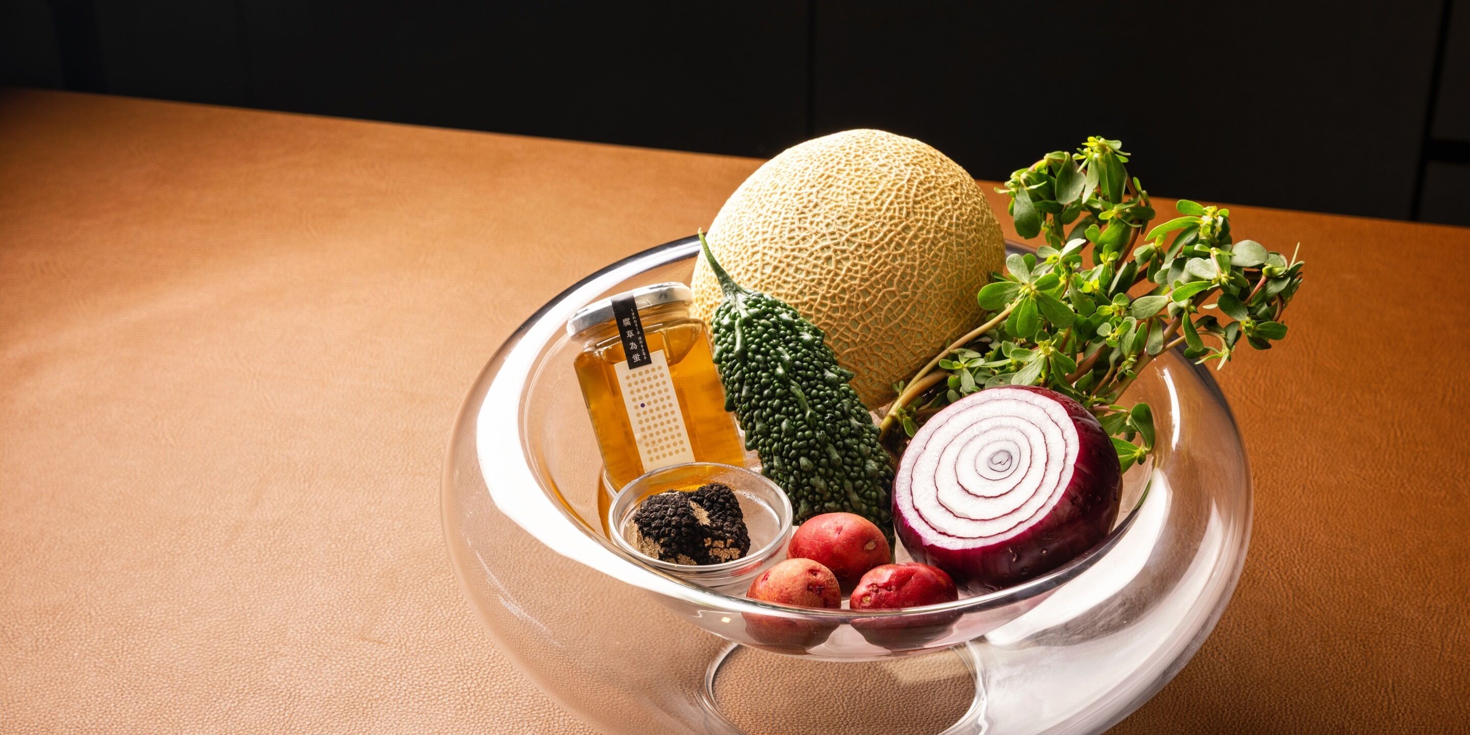 Un saladier en verre transparent contient un melon cantaloup, un oignon rouge, du poivre noir et une petite botte d’herbes aromatiques, le tout posé sur une table en bois.