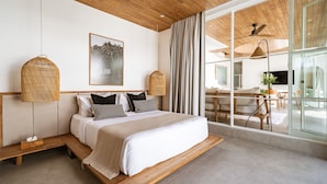 A modern bedroom with a wooden ceiling, a large bed with white bedding, a wicker lamp, and a view of an outdoor dining area through large windows.