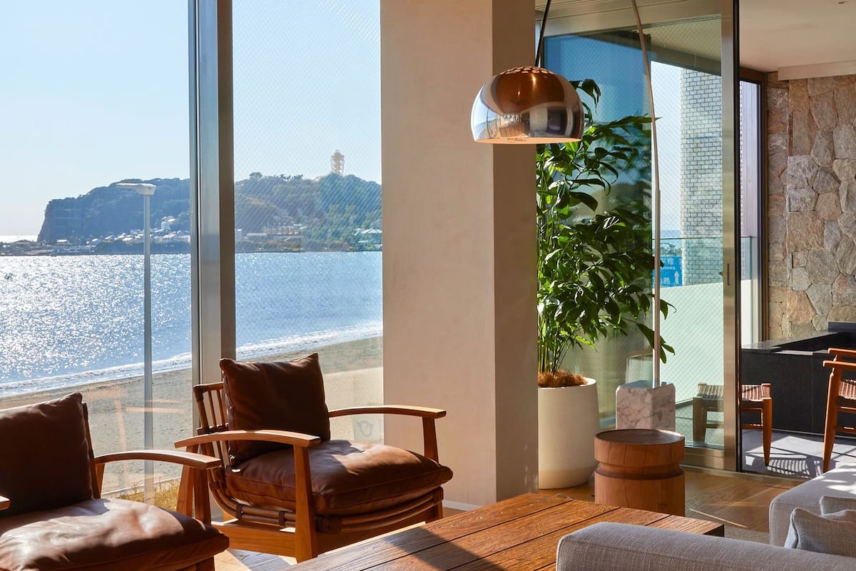 Habitación con un ventanal grande que da a una playa y una colina distante. Hay dos sillones de cuero y una mesa de madera.