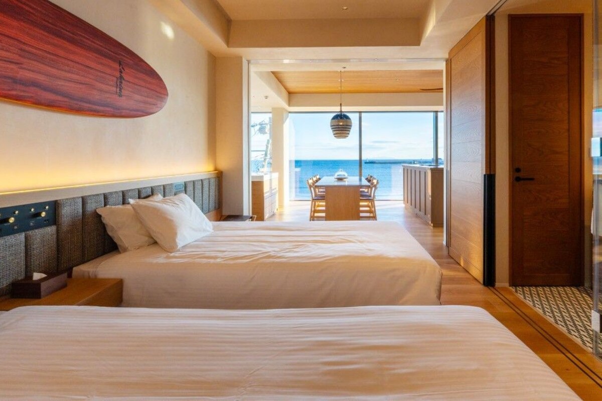 A hotel room with two beds, a view of the sea, a dining area, and a glass door leading to a balcony.