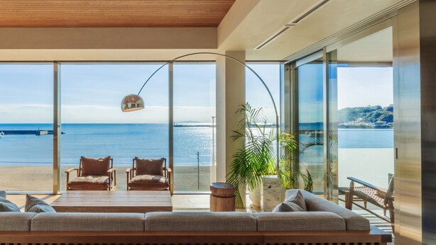 A modern living room with a large glass wall overlooking a beach and ocean. There are comfortable seating areas, a coffee table, and a lamp. - UMITO KAMAKURA KOSHIGOE (Kamakura)
