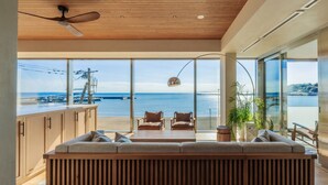 A modern living room with a large window overlooking a beach and the sea. There is a wooden ceiling fan, a sofa, and a dining table. - UMITO KAMAKURA KOSHIGOE (Kamakura)