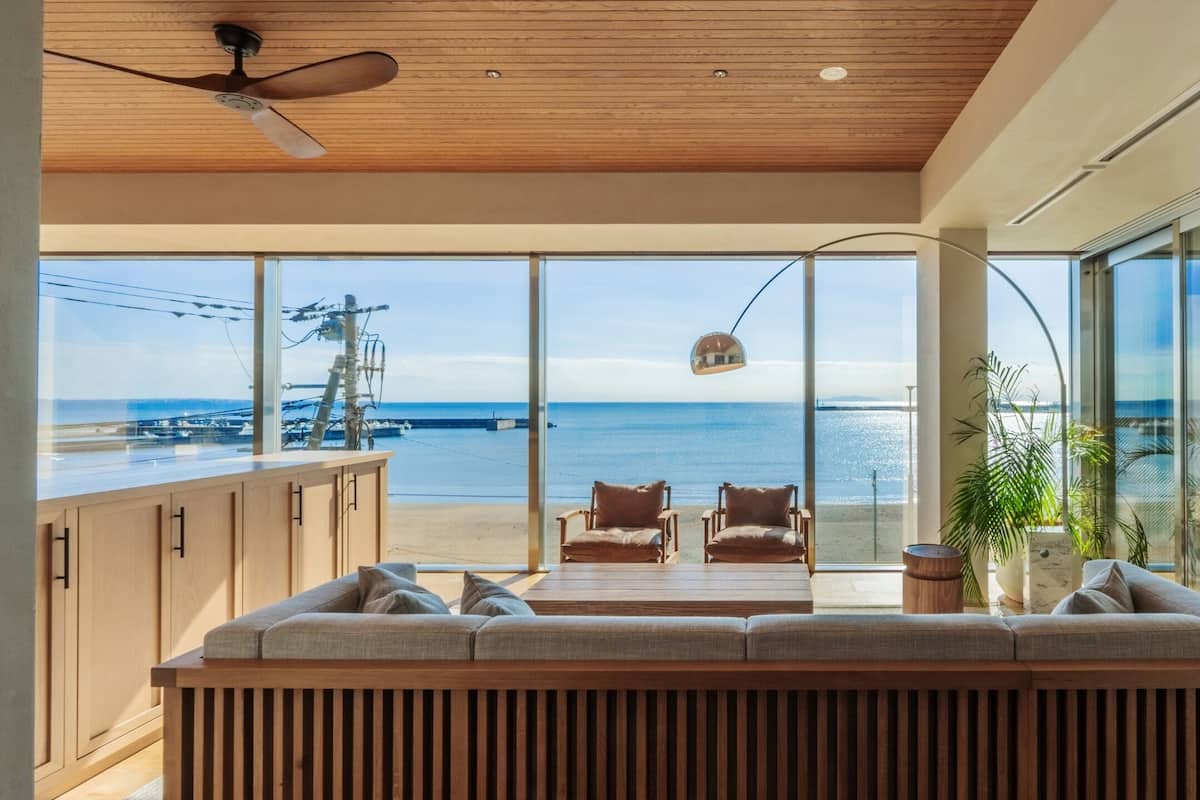 Una sala de estar moderna con un amplio ventanal que da a una playa y al mar. Hay un ventilador de techo de madera, un sofá y una mesa de comedor.
