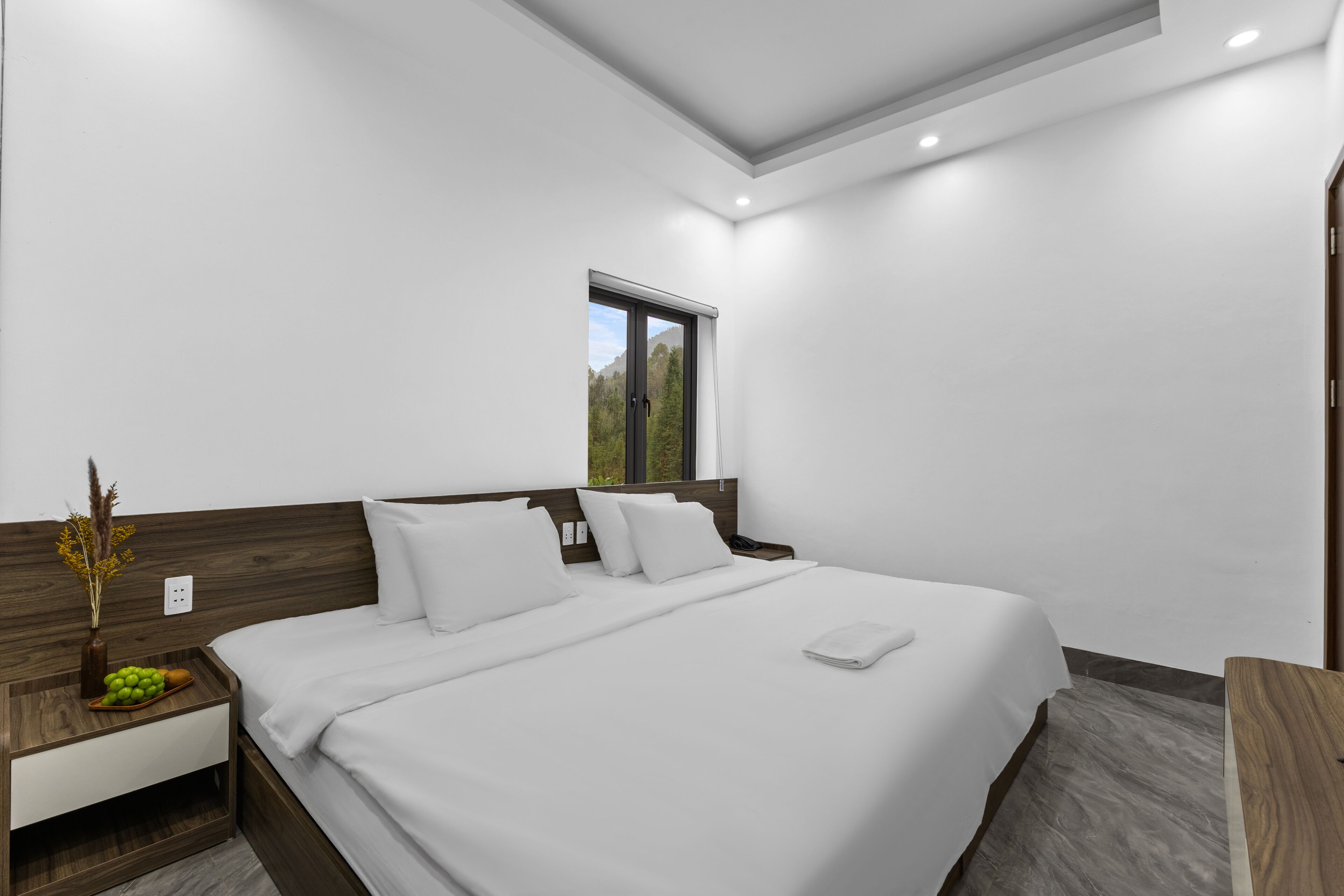 A modern hotel room with a large bed, a nightstand, a window, and a wooden headboard.