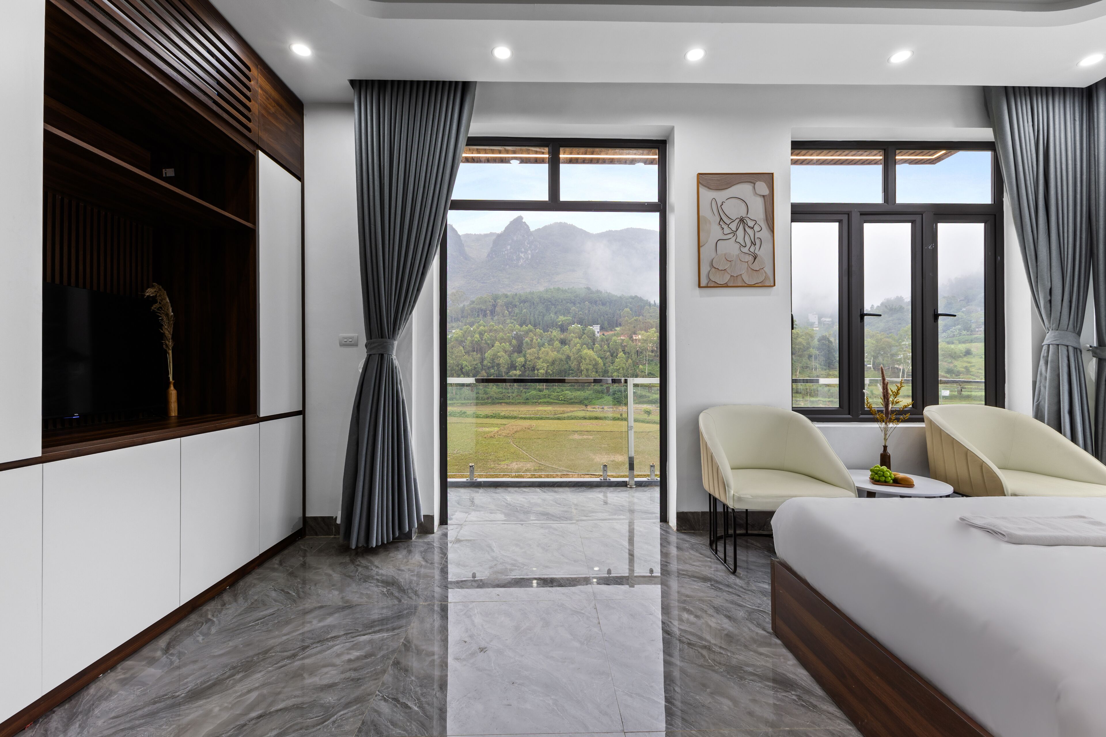 Habitación de hotel moderna con una cama grande, dos sillas, una mesita y un balcón con vistas a la montaña.