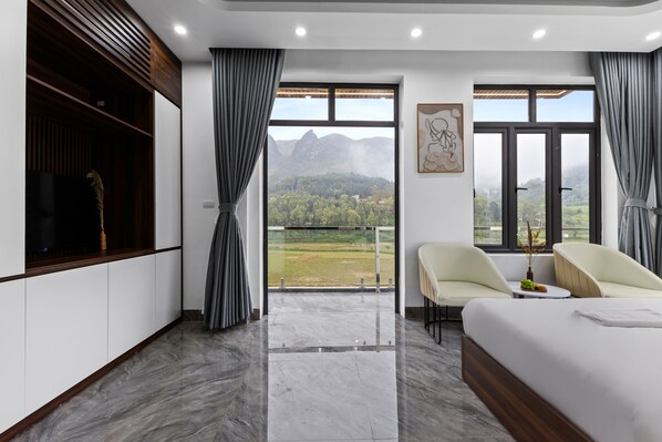 A modern hotel room with a large bed, two chairs, a small table, and a balcony with a mountain view.
