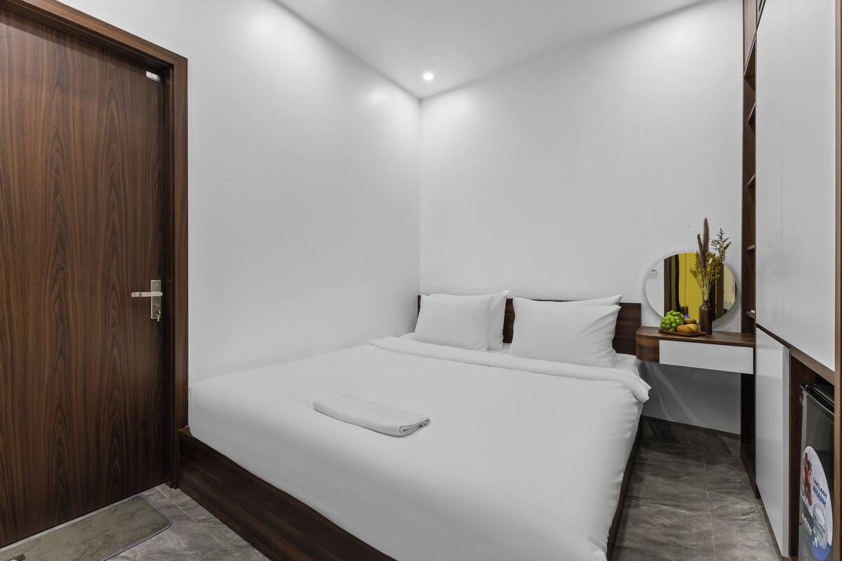 Quarto de hotel com porta de madeira, cama com roupa de cama branca, mesa de cabeceira com um vaso e um pequeno espelho.