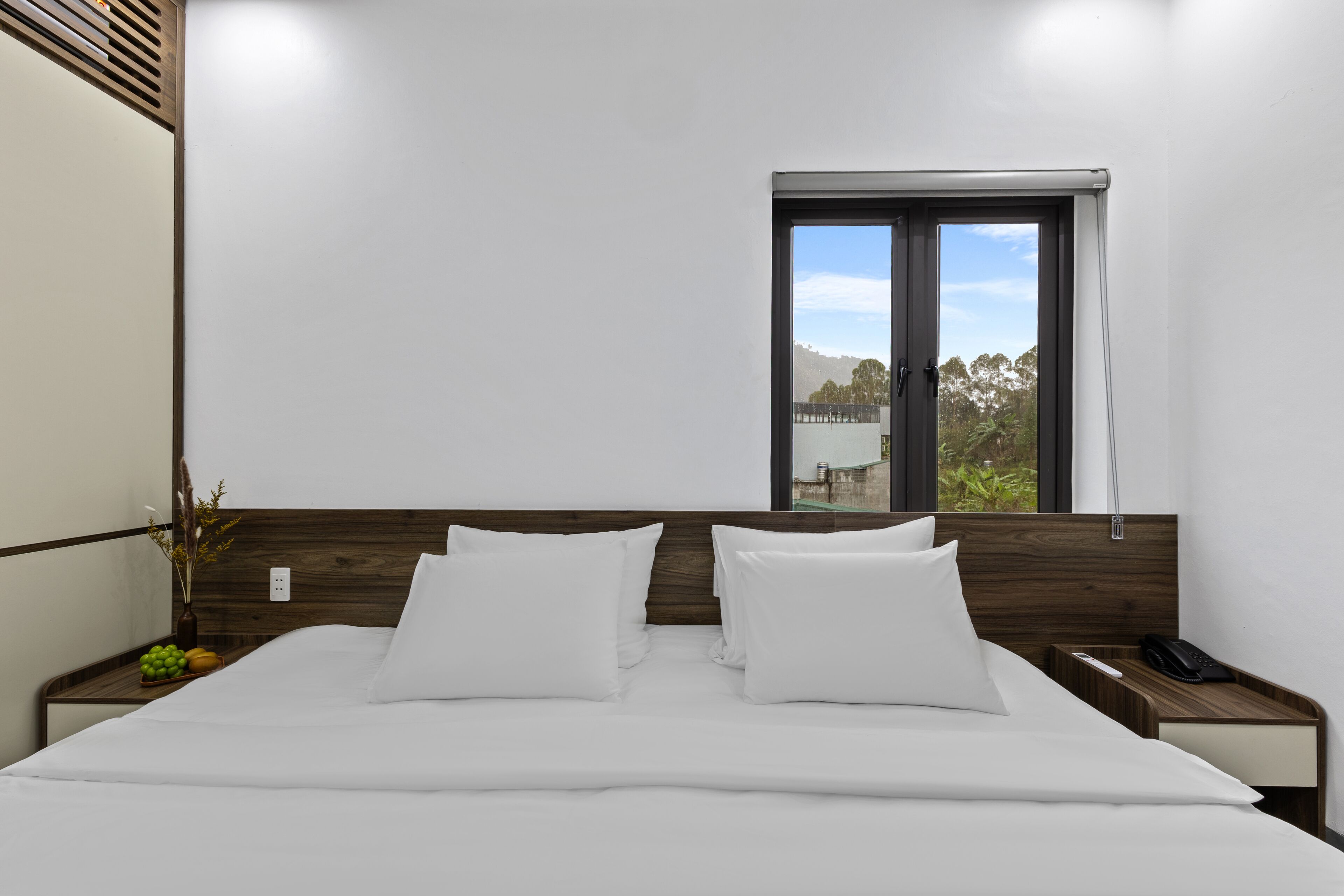 Cama bem arrumada com lençóis e almofadas brancas, cabeceira de madeira, mesa de cabeceira com telefone e janela com vista para vegetação e um edifício.